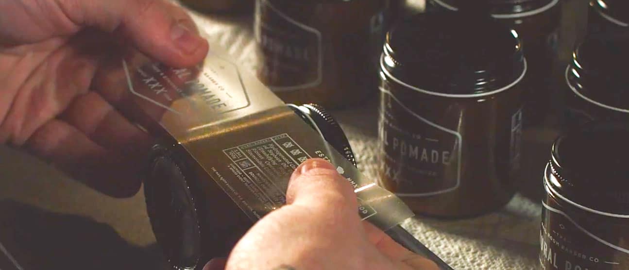 Person applying clear labels to a jar of pomade.