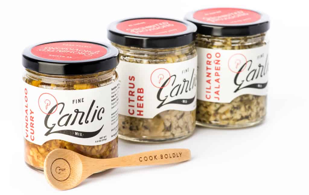 Three jars of Prohibited Provisions artisan garlic with a specialty wooden spoon.